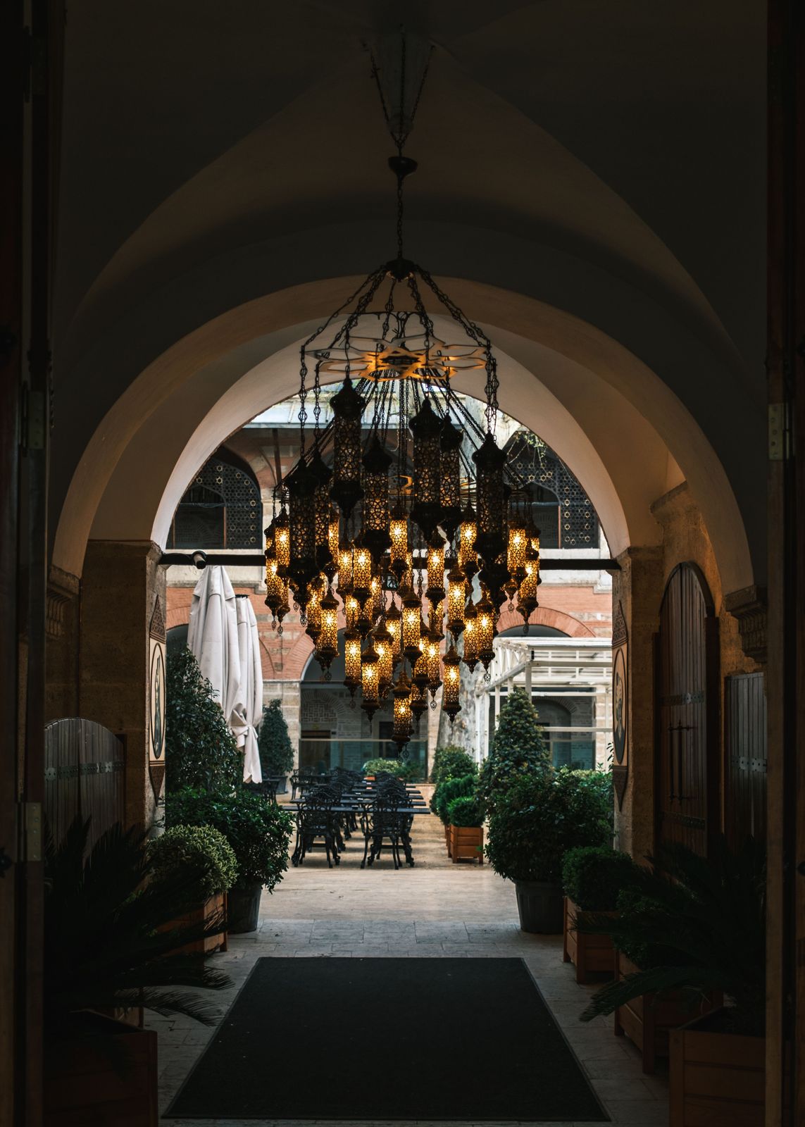 Chandelier made of dozens of pierced-metal cylinder lanterns suspended from a star-shaped frame inside a stone archway.