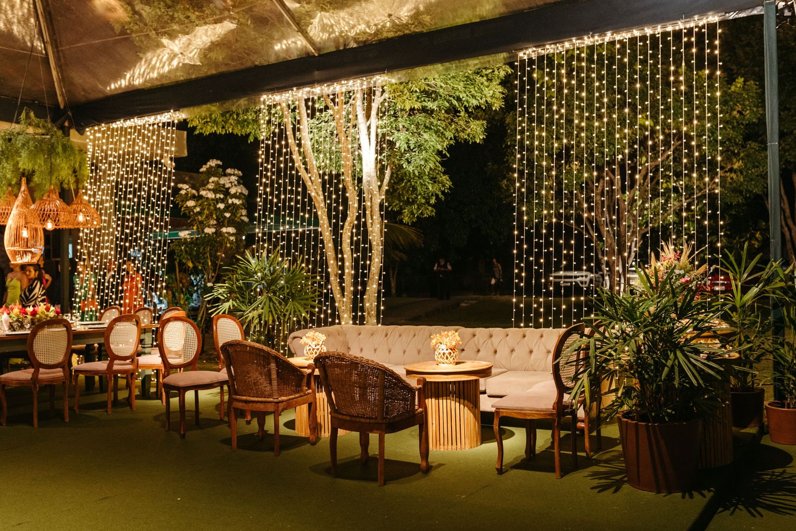 Vertical curtain of warm-white string lights draped from a tent edge behind outdoor lounge furniture and trees.