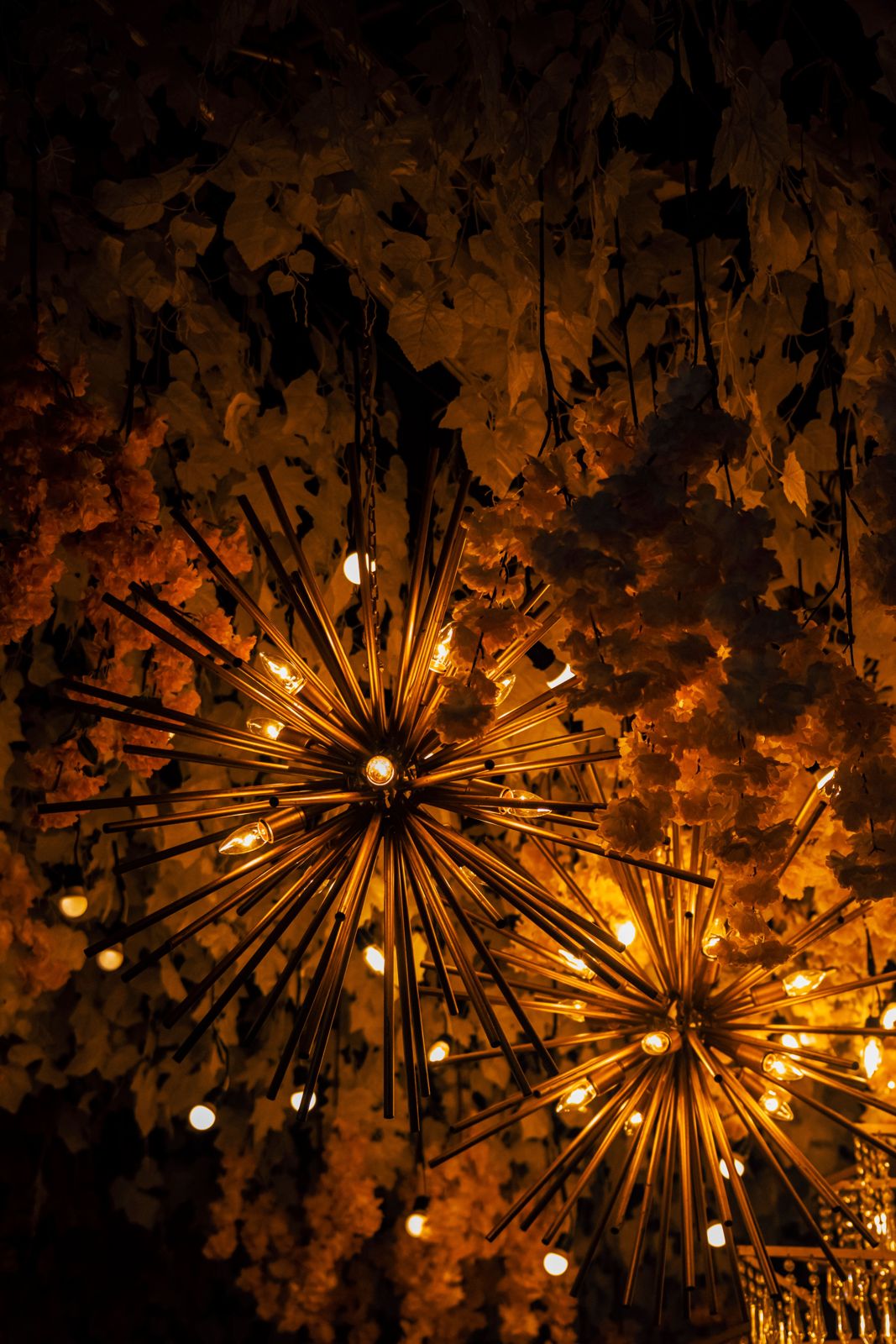 Gold sunburst sputnik pendant light with radiating rods and candle bulbs set against hanging flowers and leaves.